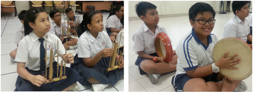 Students in school uniforms sit on the floor holding traditional instruments including bamboo xylophones and tambourines.